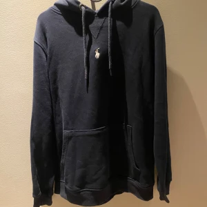Marinblå  hoodie från Polo Ralph Lauren - Marinblå hoodie från Polo Ralph Lauren med klassisk huva och snörning. Tröjan har en broderad logga på bröstet och en stor magficka framtill. Mjuk bomullskvalitet och långa ärmar gör den perfekt för chill dagar. Jag är 180 lång och den sitter fint.