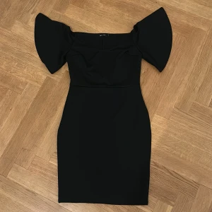 Svart offshoulder klänning NLY ONE - Supersnygg svart kortklänning från NLY Eve med offshoulder och vida, dramatiska ärmar. Figurnära passform som framhäver siluetten och ger en elegant look. Perfekt för dig som vill sticka ut med stilrena detaljer och modern design.
