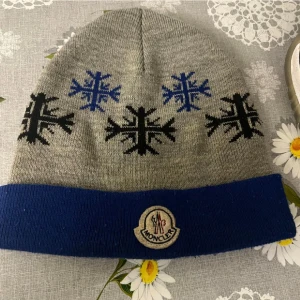  Moncler mössa  - Snygg grå mössa från Moncler med blå uppvikt kant och broderad logotyp framtill. Mössan har stickat mönster med svarta och blå snöflingor runtom. Perfekt för kalla dagar och en stilren vinterlook.