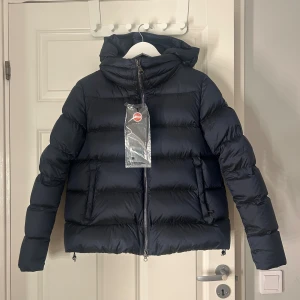 Mörkblå Colmar pufferjacka med huva - Snygg mörkblå pufferjacka från Colmar med huva och tvåvägsdragkedja. Jackan har en quiltad design, två sidofickor med dragkedja och en klassisk logga på ärmen. Det finns även en dragkedja som ger möjlighet att ta av luvan. Perfekt för kalla dagar och riktigt skön att bära. Helt ny ändats testad!