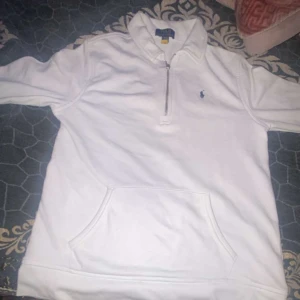 Vit half zip tröja Polo Ralph Lauren - Snygg vit långärmad tröja från Polo Ralph Lauren med half zip-dragkedja och klassisk krage. Tröjan har en broderad blå logga på bröstet och en stor magficka framtill. Tillverkad i mjuk bomull, perfekt för chill dagar.