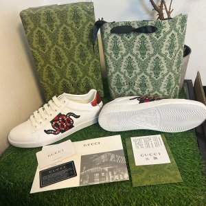 Gucci Ace sneakers med orm och ränder - Gucci Ace sneakers i vitt skinn med ikonisk röd och grön orm-broderi på ena sidan och klassiska gröna/röda ränder på andra. Baksidan har metallicröd och metallicgrön detalj med Gucci-logga. Platt sula, snörning och rund tå. Snygg och lyxig design som sticker ut.