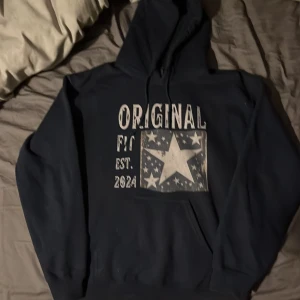 Mörkblå hoodie med stjärntryck Gildan - Snygg mörkblå hoodie från Gildan med stort tryck på bröstet där det står 'ORIGINAL' och en stor vit stjärna omgiven av mindre stjärnor. Tröjan har huva med snörning och en klassisk känguruficka framtill. Perfekt för dig som gillar streetwear vibes.