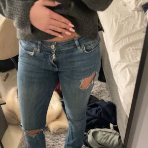 Blå bootcut jeans med slitningar - Säljer ett par blå jeans med bootcut passform och snygga slitningar vid knäna. Jeansen har klassisk femficksdesign, knapp och dragkedja framtill samt låg midja. Perfekta för dig som gillar en avslappnad och trendig stil.