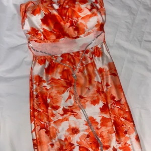 Figursydd klänning - Sjukt snygg klänning med orangea blommor. Klänningen har inbyggd bh, är axelbandslös, är öppen under brösten där lite av magen visas, med en silvrig detalj samt slits nedtill. Klänningen når till ovanför knäna. Den sitter tight och är figursydd i stretchigt material 💞Storlek S