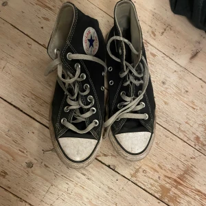 Svarta Converse Chuck Taylor High - Klassiska svarta Converse high-top sneakers i storlek 38 med vit gummitå och sula. Skorna har grå snörning, vit kontrastsöm och rund tå. Märkets logga på sidan och slitstark canvas i ovandelen. Perfekt för dig som gillar ikonisk streetstil. Den är dock lite trasig inuti vid hälen jag kan skicka bild om behov!💕