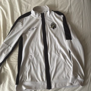 AIK Tröja Nike S/M - Snygg vit och svart vindjacka från Nike med AIK-emblem på bröstet och tryck med årtalet 1891 på ryggen. Jackan har dragkedja framtill, två fickor och är tillverkad i Dri-Fit polyester. Storlek S men passar mer åt M-hållet.