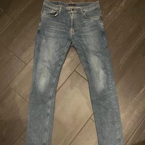 Blå jeans från Nudie Jeans W33 L34 - Säljer ett par klassiska blå jeans från Nudie Jeans i storlek W33 L34. Jeansen har en rak passform med snyggt slitna detaljer och orangea sömmar. Tillverkade i bomull med fem fickor och dragkedjegylf. 