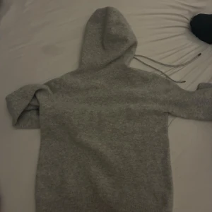 Grå stickad Kaschmir hoodie  - En fin Kaschmir grå hoodie med huva och snörning, tillverkad i stickat material. Tröjan har långa ärmar och ribbade muddar vid ärmslut och nederkant. Perfekt för dig som gillar enkel och chill stil.