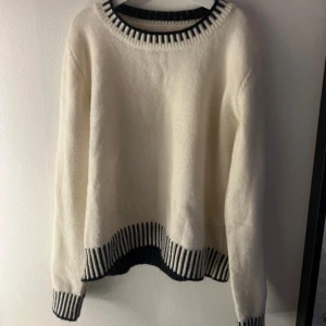 Vit stickad tröja med svarta detaljer - Snygg vit stickad tröja med svarta, randiga ribbade detaljer vid halsringning, nederkant och ärmslut. Tröjan har en rund hals och långärmade ärmar, perfekt för att hålla dig varm och stilren under kyliga dagar.