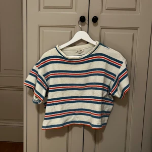 Randig croppad topp från We are Kids - Säljer en croppad topp från We are Kids i storlek XS/S. Toppen är vit med blå och röda ränder, har korta ärmar och rund hals. Materialet är frotté vilket gör den extra mjuk och chill. Perfekt för dig som gillar retrovibbar och färg!