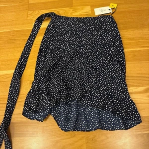 Blå prickig omlottkjol Amelie & Me - Supersöt blå omlottkjol från Amelie & Me med vita prickar och volangkant nertill. Kjolen har knytband i midjan och är i ett lätt och luftigt material, perfekt för varmare dagar. Ny med prislapp. 