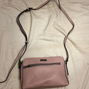 Rosa axelväska från Don Donna - Snygg ljusrosa axelväska från Don Donna i skinnimitation med flera fack och dragkedjor. Väskan har justerbar axelrem och silverfärgad logga framtill. Perfekt storlek för det viktigaste och stilren design med svarta detaljer längs kanterna.