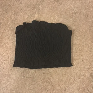 Svart bandeau tubtopp från SHEIN - Svart bandeau tubtopp från SHEIN i storlek XS Petite. Toppen är smockad med volangkant upptill och nertill, vilket ger en snygg och trendig look. Perfekt att matcha med höga jeans eller kjol.