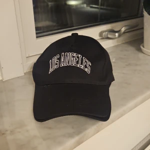 Svart keps med Los Angeles-tryck - Svart keps med böjd skärm och vit broderad text 'LOS ANGELES' framtill. Justerbar rem med metallspänne baktill för perfekt passform. Snygg och enkel design som passar till streetwear och casual outfits. Materialet är bomull och kepsen har en klassisk form.