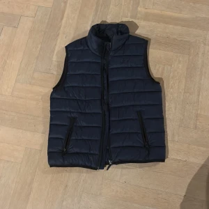 Mörkblå dunväst från Kappahl - Snygg mörkblå dunväst från Kappahl i storlek 158. Västen har quiltad design, hög krage och två praktiska fickor med dragkedja. Perfekt att slänga över en hoodie för extra värme. Stängs med dragkedja framtill och har en stilren look. Den är knappt använd, nästan helt ny.