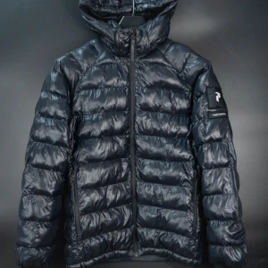 Svart Peak Performance dunjacka 170 - Snygg svart dunjacka från Peak Performance med glansig finish och huva. Jackan har lång ärm, dragkedja framtill och logotyp på vänster ärm. Perfekt för kalla dagar och har en quiltad design som ger extra värme och stil. Storlek 170