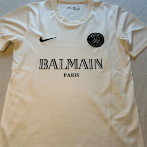 Vit PSG x Balmain t-shirt Nike - Snygg vit t-shirt  Nike med PSG och Balmain Paris tryck på bröstet. Klassisk rund halsringning och korta ärmar. Tillverkad i 100% polyester med Dri-FIT-teknologi för extra komfort. Perfekt för dig som gillar streetwear och sportig stil.