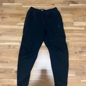 Svarta Nike tech mjukisbyxor - Svarta Nike tech mjukisbyxor från Nike med dragsko i midjan och två sidofickor. Byxorna har en slim passform och är tillverkade i mjukt bomullsmaterial. Nike-logga i svart på vänster ben ger en clean och sportig look. Storlek S, knappt använda och inga defekter eller sådant! 