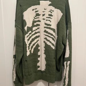 Grön stickad tröja från Kapital - Cool grön stickad tröja med vit skelettdesign på baksida samt ärmar. Tröjan har en loose passform, ribbade muddar och långärmade. Perfekt för dig som gillar edgy och unik stil. Storlek L