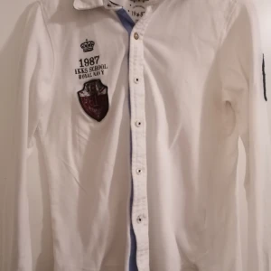 Vit långärmad skjorta från IKKS - Snygg vit skjorta från IKKS med broderade detaljer och patch på bröstet, samt stort svart nummer på ärmen. Skjortan har klassisk krage, knappar framtill och coola inslag av blått tyg vid kragen. Perfekt för dig som gillar preppy stil med unika detaljer.