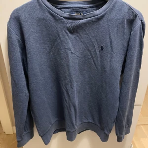 Blå sweatshirt från Polo Ralph Lauren - Blå sweatshirt från Polo Ralph Lauren med klassisk rund hals och broderad logga på bröstet. Tröjan har långa ärmar och ribbade muddar vid ärmslut och nederkant. Perfekt för chill dagar och enkel att matcha med jeans eller joggers.