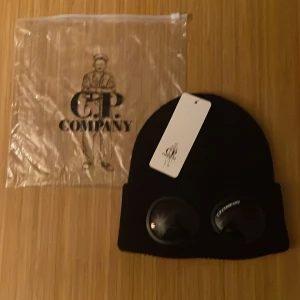 Svart C.P. Company mössa med goggles - Svart mössa från C.P. Company med ikoniska goggles på uppviket. Stilren och urban design, perfekt för dig som vill sticka ut. Mössan är ribbstickad och har en klassisk form med rundad topp. Materialet är mjukt och värmande, och loggan syns tydligt på ena glaset. Helt ny!!!