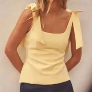 Ljusgul linnetopp med rosettband - Supersöt ljusgul linnetopp med breda axelband som knyts till rosetter på axlarna. Toppen har en fyrkantig urringning och är figurnära med en stilren, enkel design. Perfekt för varma dagar och ger en fräsch vibe till din outfit.