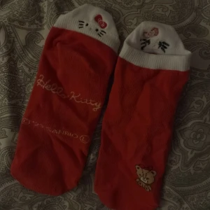 Hello Kitty strumpor i rött och vitt - Säljer ett par söta Hello Kitty strumpor från Sanrio i rött och vitt. Strumporna har Hello Kitty-ansikte broderat på tårna och en liten nalle med rosett på sidan. Mjuk bomullskvalitet och låg modell, perfekt för sneakers. Hello Kitty-logga broderad på ena strumpan.