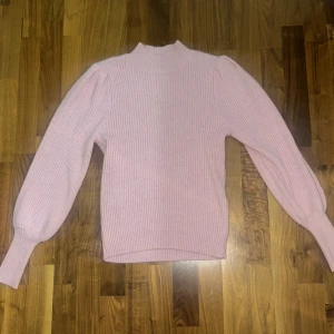 Rosa ribbad stickad tröja från ONLY -  Ljusrosa stickad tröja från ONLY med ribbat mönster och puffiga långärmar. Tröjan har en högre halsringning och är perfekt för både en uppklädd outfit men även basic. 