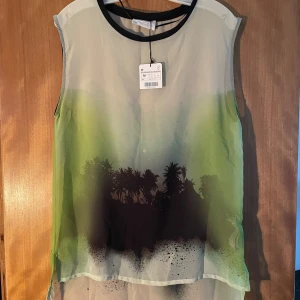 Grön och svart blus med palmmotiv - Säljer en luftig blus från Culture i transparent syntetmaterial. Blusen är ärmlös och har ett coolt palmmotiv i svart mot en grön och beige bakgrund. Rund halsringning med svart kant. Perfekt för dig som gillar tropiska vibes.