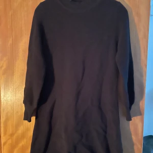 Svart stickad långärmad klänning - En svart stickad klänning med lång ärm och rund hals. Modellen är enkel och stilren, perfekt för dig som gillar en klassisk look. Materialet är mjukt och ger en skön känsla mot huden. Passformen är normal och klänningen har en kort längd.