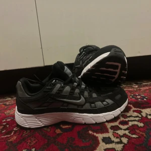 Nike sneakers svart och grå mesh - Nike sneakers i svart och grå med vit sula. Ovandelen är i mesh och syntet med coola detaljer och snörning. Swoosh-logga på sidan och Nike-märke på plösen. Skorna har en sportig vibe och passar perfekt till streetwear.