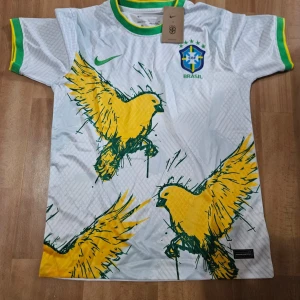 Vit Brasilien fotbollströja Nike S - Snygg vit Brasilien fotbollströja från Nike med gröna och gula detaljer på krage och ärmslut. Framsidan har stora gula fåglar och CBF-emblem broderat på bröstet. Tillverkad i Dri-FIT-material som andas, perfekt för match eller träning.