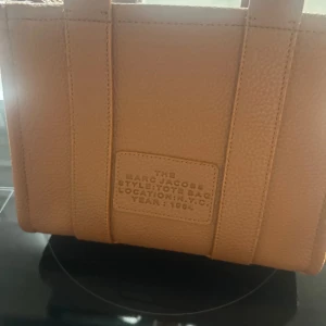 Beige Tote Bag från Marc Jacobs - Snygg beige Tote Bag från Marc Jacobs i strukturerat skinn. Väskan har tydlig logga framtill, robusta handtag och dragkedja upptill. Insidan har ett fack med dragkedja och detaljer i samma färg. Perfekt för dig som vill ha en trendig och rymlig väska.