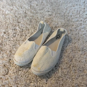 Vita ballerinaskor med broderade blommor - Säljer ett par vita ballerinaskor från Clou i storlek 38. Skorna har ett broderat blommönster över hela ovandelen och är gjorda i bomullstyg med platt sula. Perfekta för vår och sommar, med en enkel och söt design.
