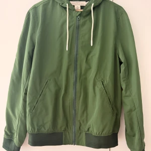 Grön vindjacka från H&M L.O.G.G. - Snygg grön vindjacka från H&M L.O.G.G. med huva och vita snören. Jackan har dragkedja framtill, två sidofickor och ribbade muddar vid ärmslut och nederkant. Perfekt för dig som gillar enkel och clean stil.