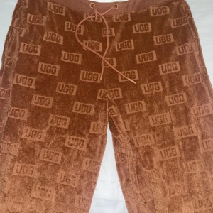 Bruna mjukisbyxor från UGG med logga - Säljer ett par bruna mjukisbyxor från UGG i storlek S. Byxorna har ett all-over UGG-logomönster, ribbad midja med snörning och mjukt frottématerial som känns riktigt skönt. Perfekt för chill dagar hemma eller när du vill vara extra comfy.