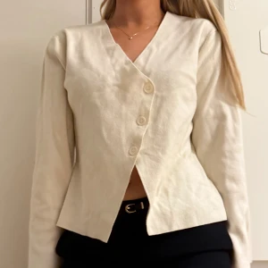 Beige kofta med sned knäppning Kappahl - Snygg beige kofta från Kappahl med unik sned knäppning framtill och tre stora knappar. Koftan har v-ringning och långa ärmar, tillverkad i mjuk bomullsmix. Perfekt för lager-på-lager och enkel att styla till olika outfits.