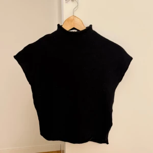 Svart polotopp i ull - Svart polotopp med korta ärmar och rak passform. Tjockt, mjukt material i ull som ger en clean och stilren look. Hög krage och croppad längd, perfekt att matcha med jeans eller kjol. Enkel design utan detaljer, passar dig som gillar minimalistiskt. Den är något krympt i tvätten.