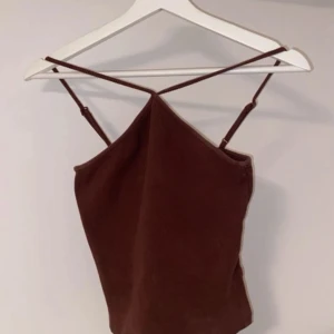 Brun croppad linnetopp med smala axelband - Säljer en brun croppad linnetopp med smala justerbara axelband som korsas i ryggen. Toppen har en enkel och stilren design med lätt A-linjeformad siluett. Perfekt för dig som gillar minimalistisk stil och vill ha något snyggt.