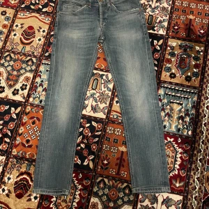 Blå Dondup Jeans i modellen George - Snygga blå jeans från Dondup med klassisk femficksdesign och diskreta slitningar. Jeansen har normalhög midja, raka ben och detaljer som bruna sömmar och metallknappar. Materialet är mjukt denim i bomull med lite stretch för extra komfort.