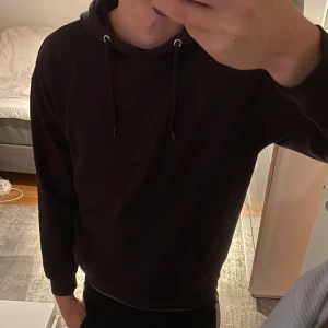 Svart hoodie med huva och ficka - Svart hoodie med klassisk huva och snörning, samt en stor magficka framtill. Tröjan har ribbade muddar vid ärmslut och nederkant, och är långärmad. Perfekt för chill dagar och enkel att matcha med jeans eller joggers.