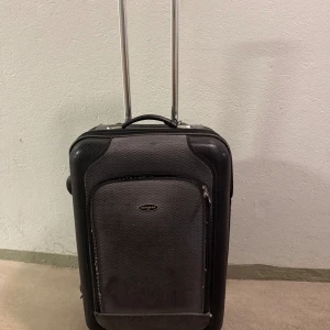 Svart resväska från Samsonite - Stilren svart resväska från Samsonite med hårt skal, teleskophandtag och praktiska hjul. Väskan har ett stort huvudfack, flera innerfickor med dragkedja och spännband för att hålla packningen på plats. Perfekt storlek för weekendresor och flyg. Ett hjul är slitet som gör att väskan rullar dåligt.