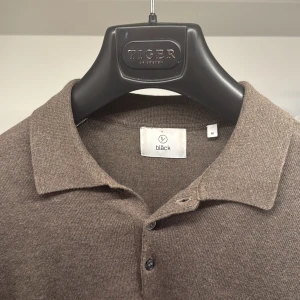 Brun pikétröja från Bläck, långärmad - Snygg brun långärmad pikétröja från Bläck med klassisk krage och två knappar framtill. Tröjan är tillverkad i mjukt stickat material som känns skönt mot huden. Perfekt för dig som gillar stilrena och enkla plagg med en chill vibe. Använd en gång! 