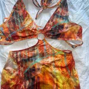 Färgstark omlottklänning med ringdetalj - Säljer en kort omlottklänning med färgstarkt abstrakt mönster i orange, gul, röd och blå toner. Klänningen har djup v-ringning, ringdetalj framtill och knyt i nacken. Modellen är tight och ärmlös, perfekt för dig som gillar att sticka ut.