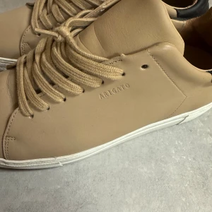 Beige sneakers från Axel Arigato - Snygga beige sneakers från Axel Arigato med stilren design och vita sulor. Skorna har snörning och är tillverkade i slätt skinn. Baktill finns svart hälparti med broderade vita tecken. Perfekt för dig som gillar minimalistisk och modern stil.