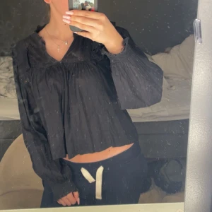 Svart blus med v-ringning och volang - Säljer en svart blus med v-ringad hals och volangdetaljer vid axlar och ärmslut. Blusen har långa, lite puffiga ärmar med smock och är croppad i modellen. Materialet är tunt och luftigt, perfekt för dig som gillar en flowy look.