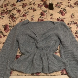 Ljusblå stickad tröja med twist - Superfin ljusblå stickad tröja med v-ringning och twistad detalj framtill. Tröjan har lång ärm och en peplum-liknande volang nertill som ger en snygg siluett. Perfekt för dig som vill ha något både mysigt och trendigt.