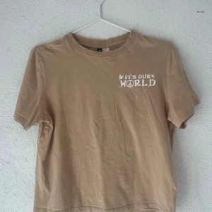 Beige t-shirt med tryck från H&M Divided - Snygg beige t-shirt från H&M Divided i 100% bomull. Framsidan har ett litet vitt tryck med texten 'IT'S OUR WORLD' och baksidan har ett stort tryck med peace-symbol, svampar och små figurer. Klassisk rund hals och korta ärmar.